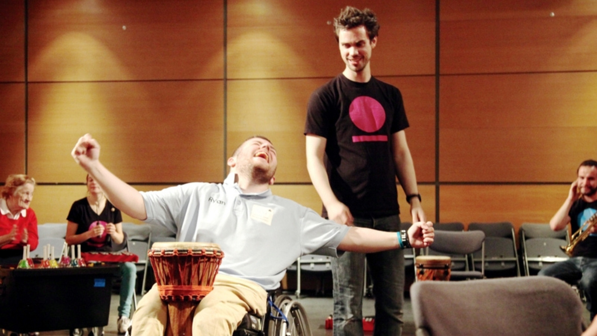 A person using a wheelchair has a drum between their knees, and their arms outstretched.