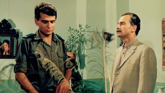 A younger man dressed in army fatigues and an older man wearing a linen suit stood in a homelike interior.