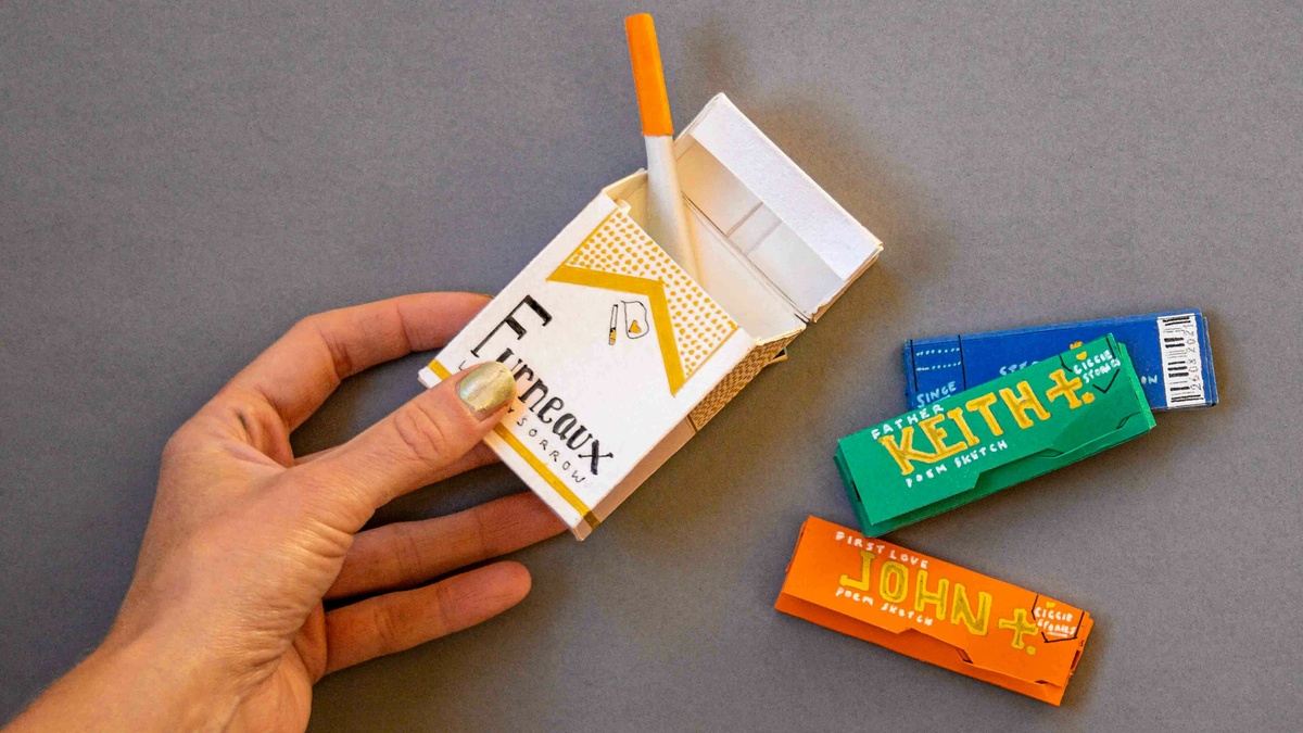 A hand holds a hand drawn packet of cigarettes made of card with the artist's surname branded on the pack.
