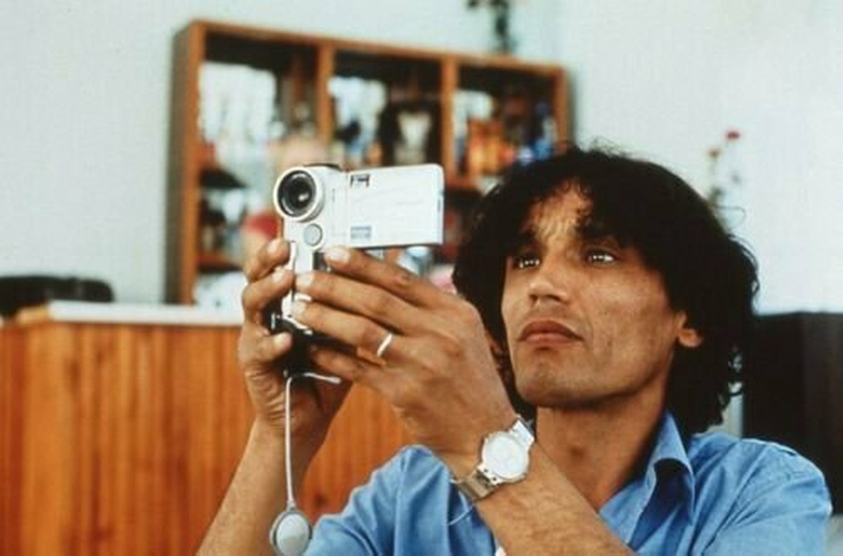 A man with curly hair in a blue shirt is holding a digital camera on his eye-level.