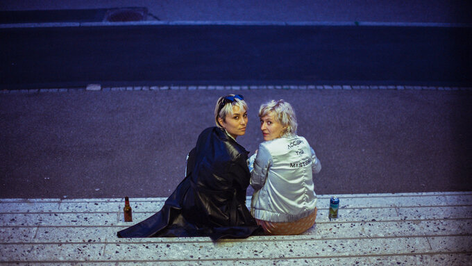 A street image with blue lighting featuring two women in the middle of the shot sitting down at some stairs.