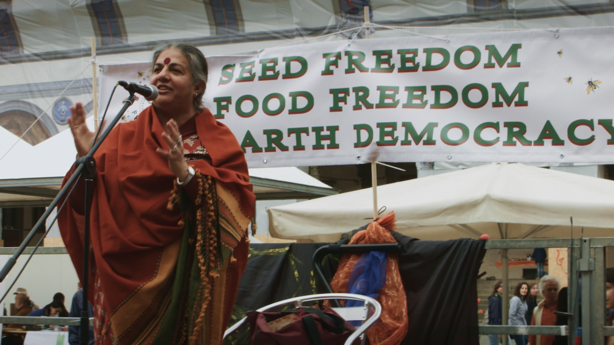 Vandana Shiva, a woman wearing a red sari and red bindi speaks into a mike before a banner which reads ‘Seed Freedom…”