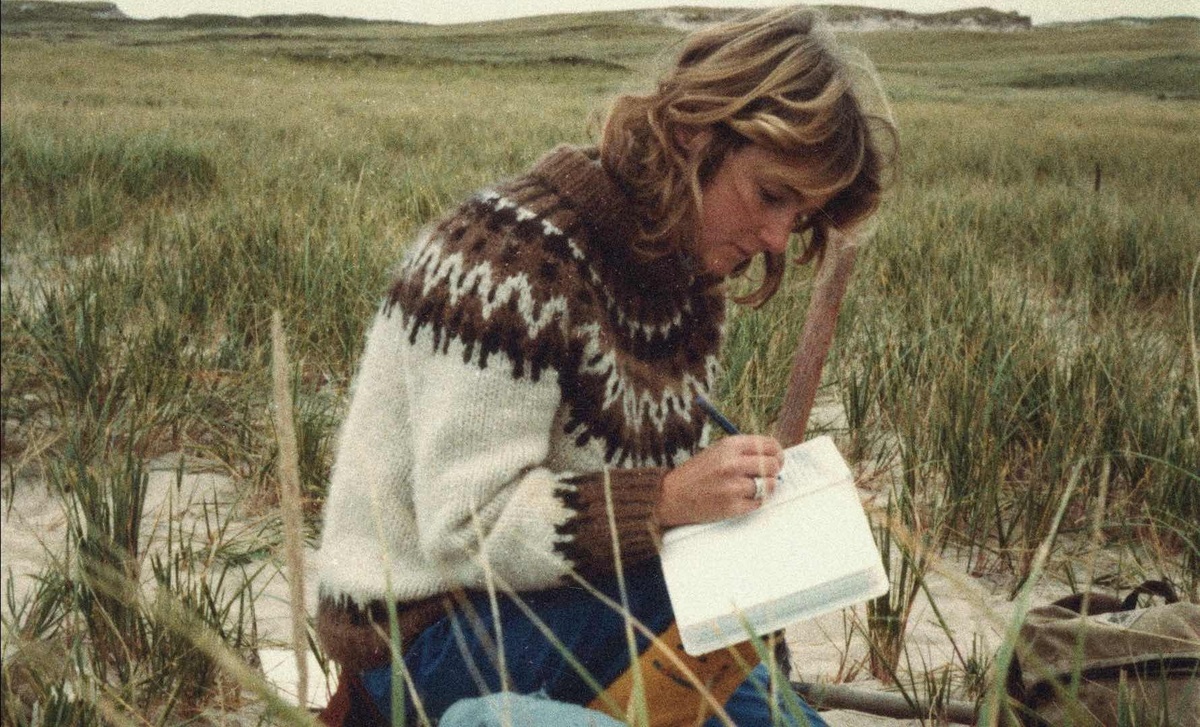 A person wearing a brown knitted jumper is sitting on a sandy and grassy landscape. They are writing in a notebook.