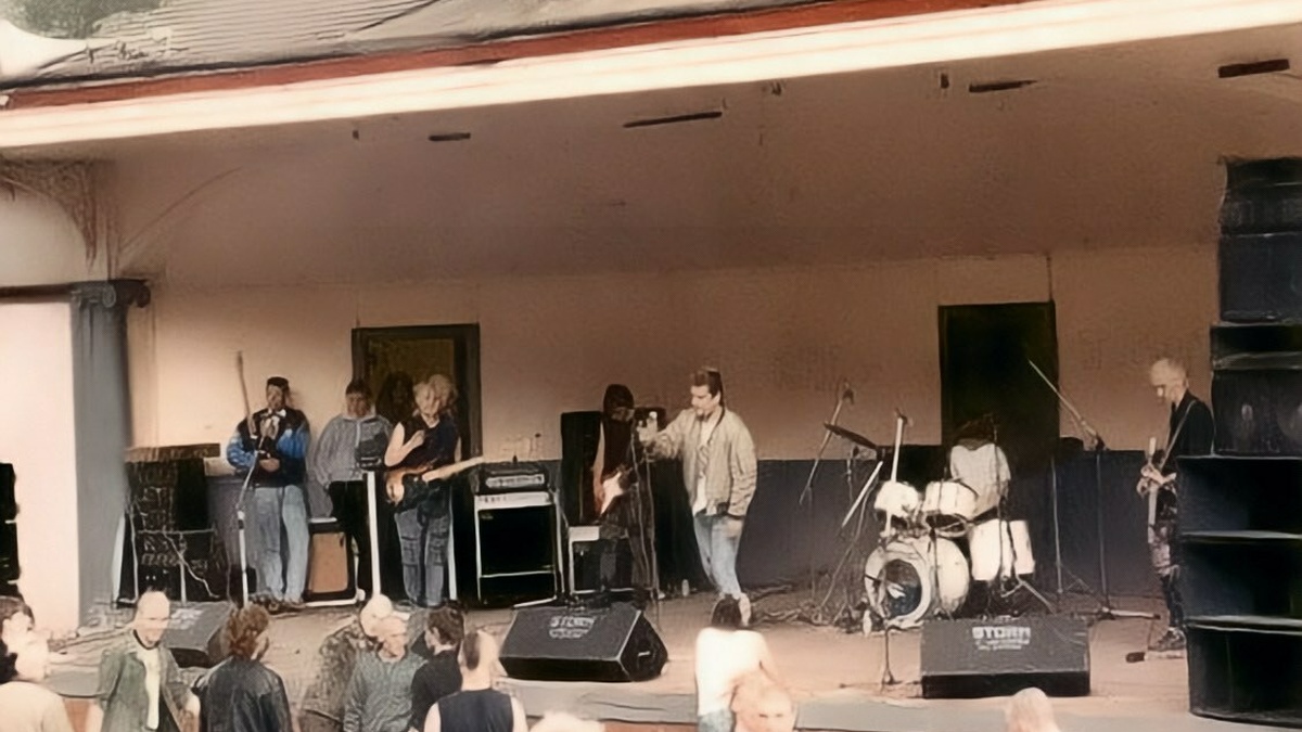 A photo of a punk band playing at an outside bandstand, a smattering of punks stand in the audience.