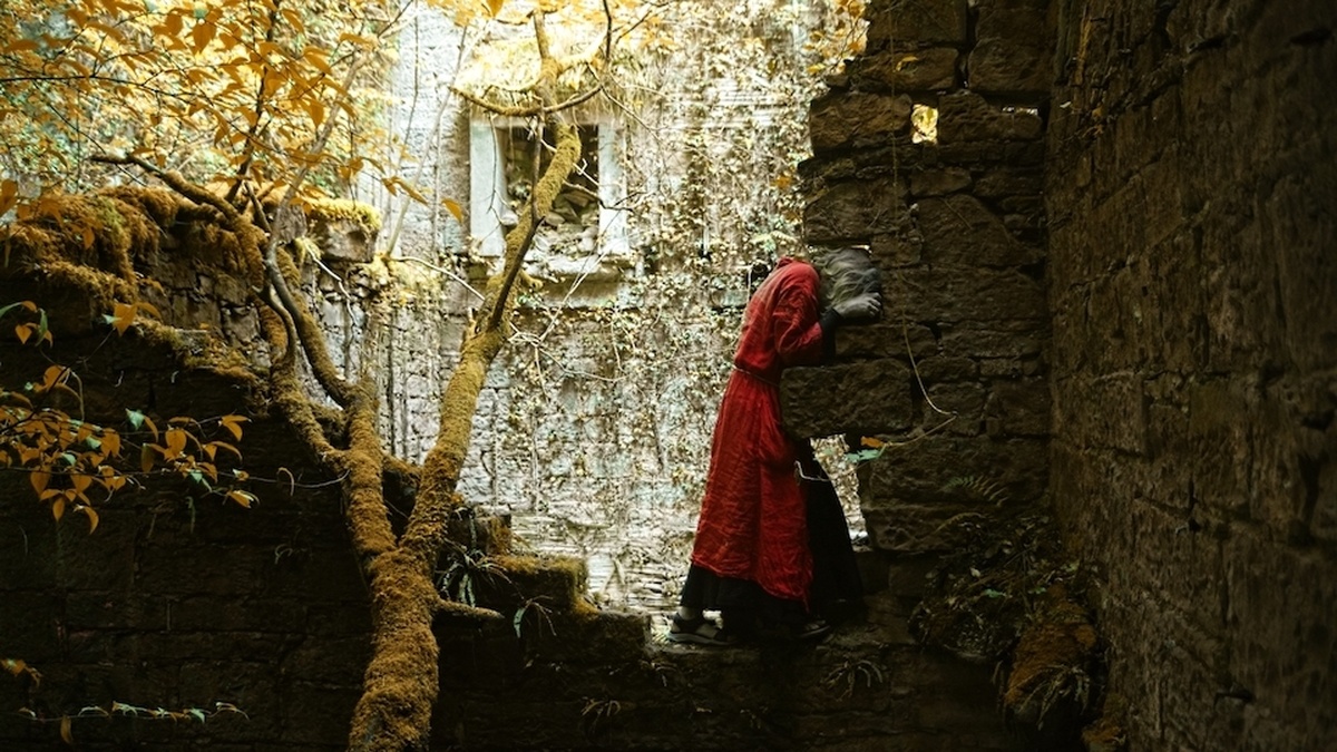 A dancer in a red gown stands amongst trees and ruins, their head inserted into a gap in the stones.