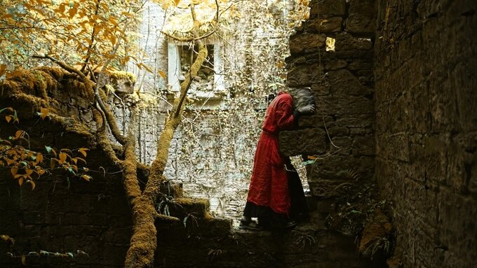 A dancer in a red gown stands amongst trees and ruins, their head inserted into a gap in the stones.
