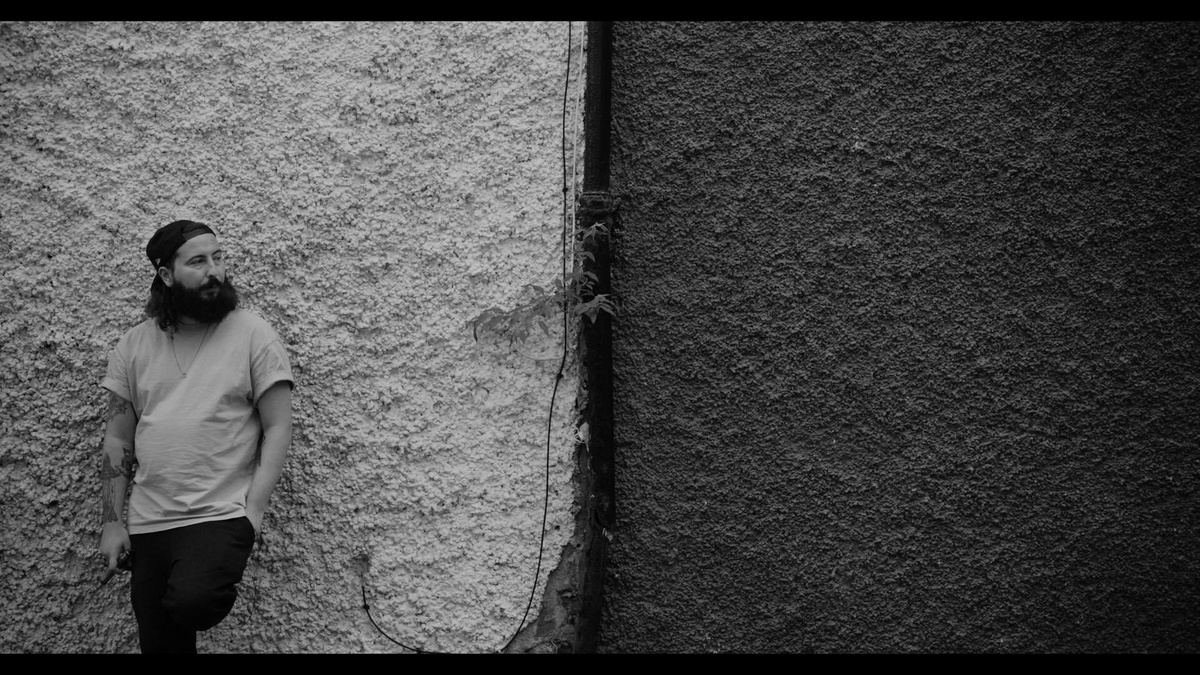 Black & White image of a bearded, tattooed young man leaning against a roughcast wall.