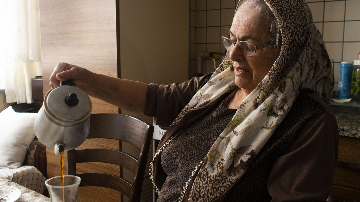 Grandmother Anik, an older woman with glasses, sits at a table, a scarf round her head, pouring tea into a glass cup.