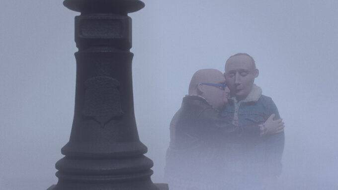 In the background of a misty day, two claymation figures embrace closely.