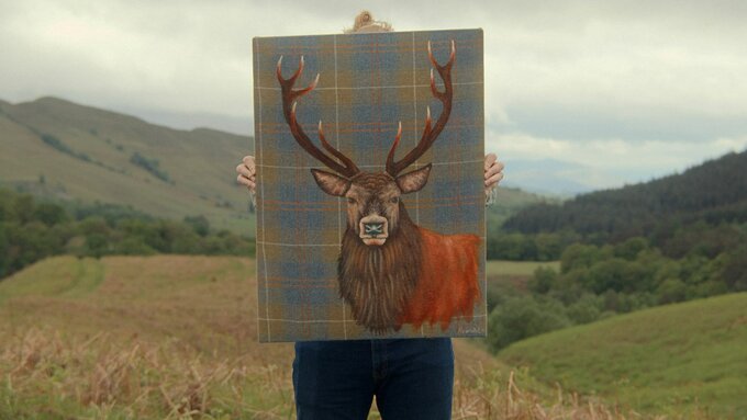 Someone holding up a painting of a deer on tartan. They are standing in front of a Highland landscape.