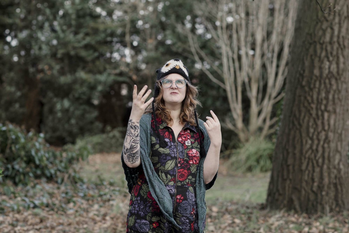 A woman with long hair in a wooded area speaking, she wears a dress, an owl beanie, & a shawl.