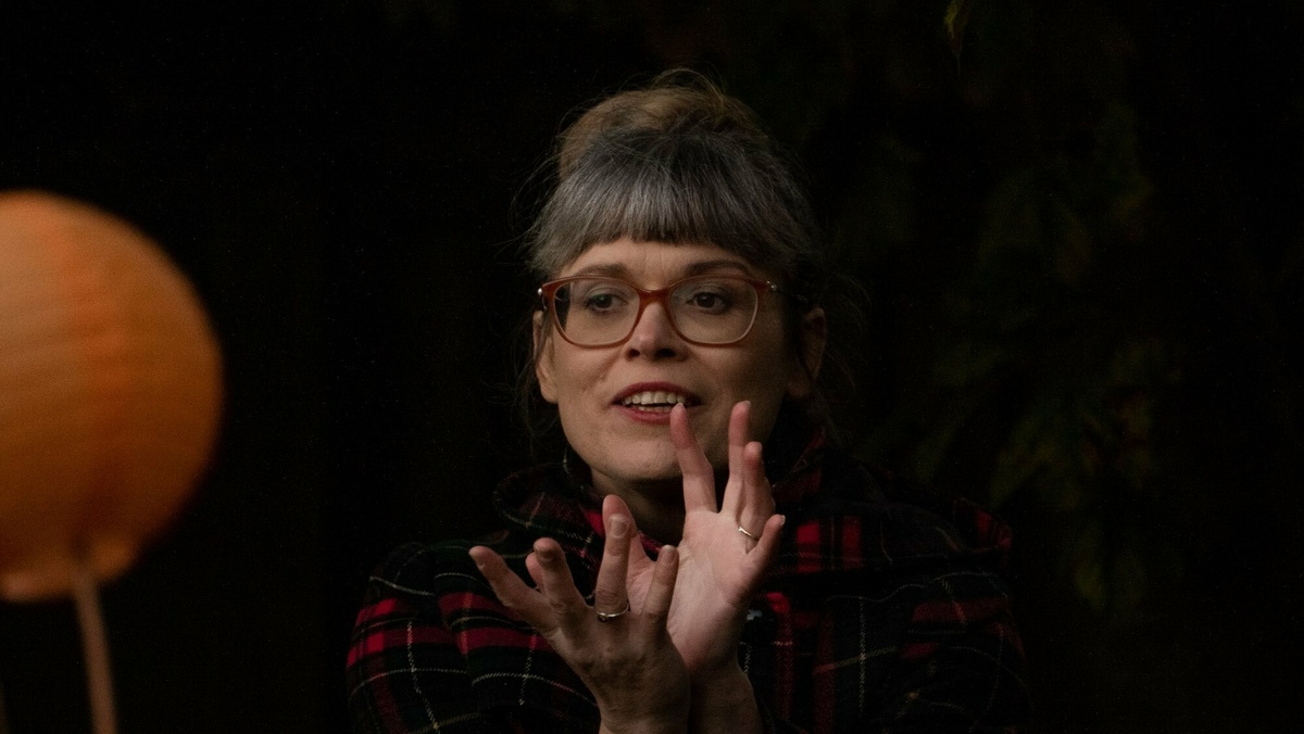 A close up image of a woman with gray hair & large glasses, her hands are held close to her face in a cryptic gesture.
