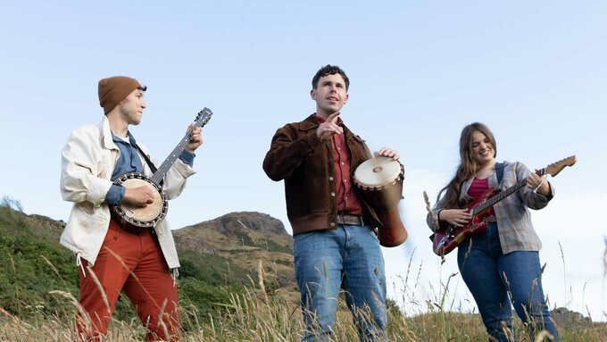 Three young people in a field on a sunny day with instruments. A banjo, a hand drum, and an electric guitar.