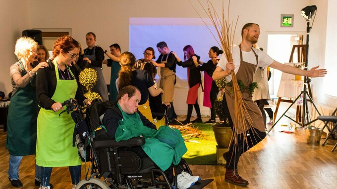 A man holding branches marches in a circle, directly behind him is a wheelchair user, part of the conga line behind him.