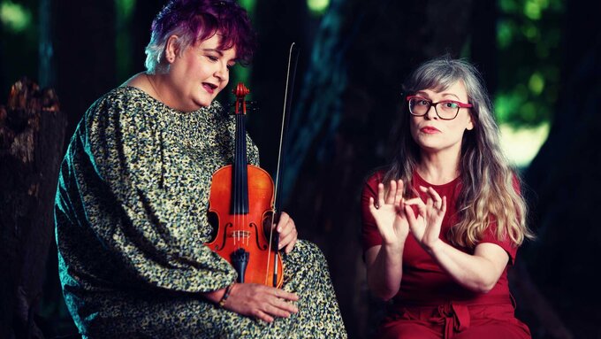 Two women in the woods. 1 holds a violin and has short purple hair. The other has long gray hair and wears a red dress.