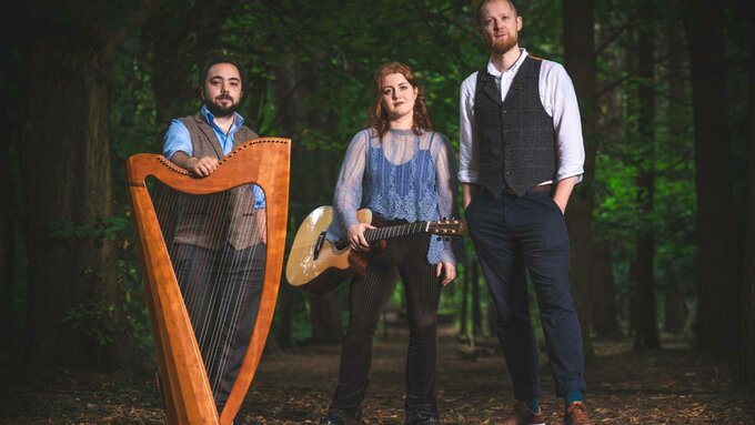 Three formally dressed people standing in the woods two with musical instruments, a large harp and a guitar.