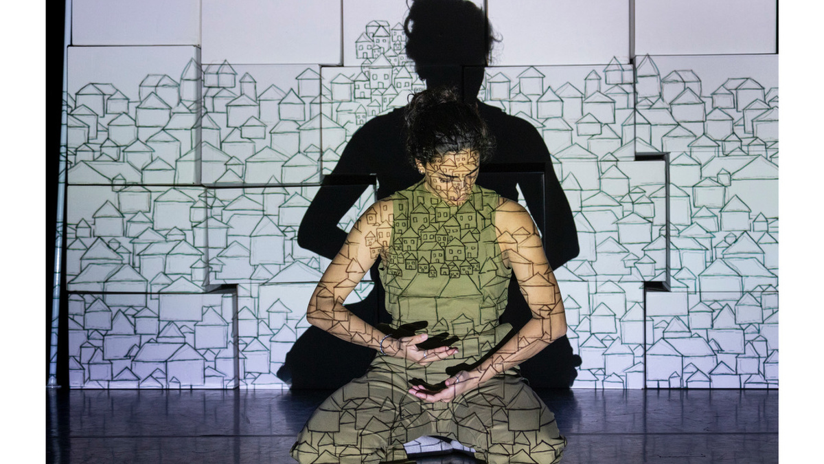 A brown skinned person wearing green sat cross legged on stage, with a drawing of houses projected onto them.