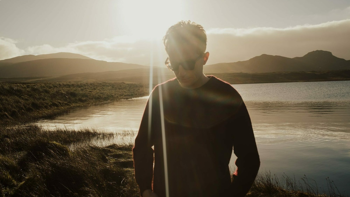 A man in sunglasses stood on the coastline as the sun sets behind him.