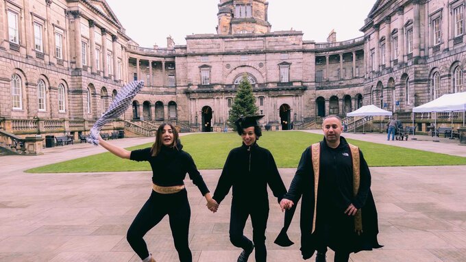Three formally dressed brown skinned people posing in part of Glasgow University, one wears a graduation cap.