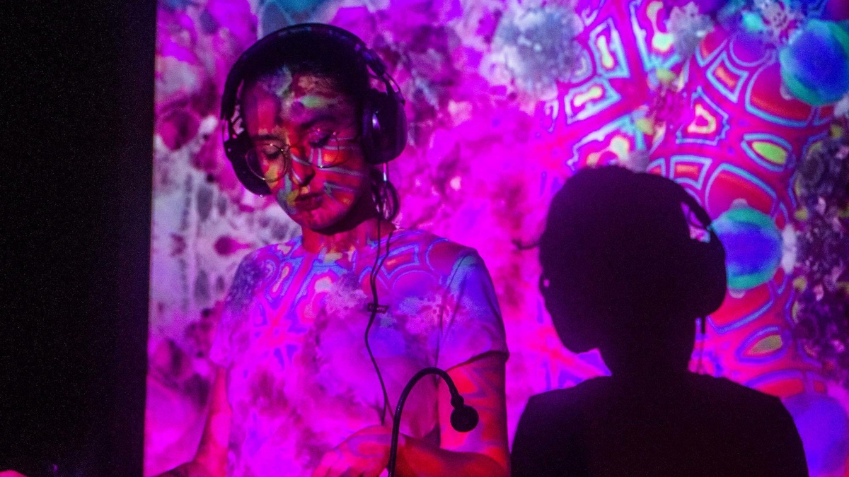 A photo of a DJ at their decks, they are lit by a pink and blue projection.