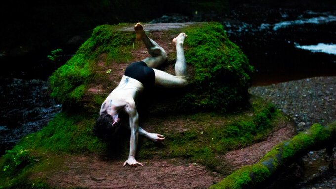 A dancer in trunks is upside down on a rocky outcrop with green moss and a river surrounding.