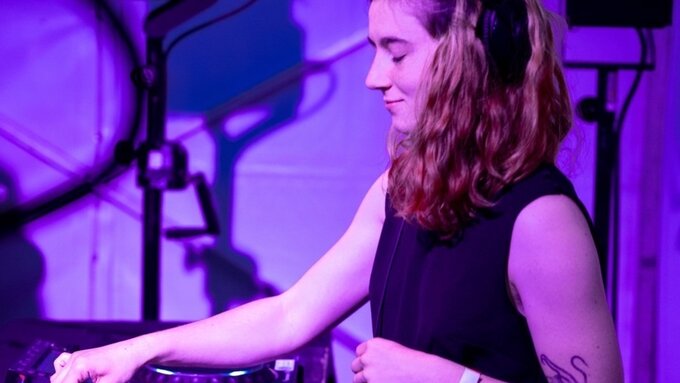 A femme person illuminated by purple light mid DJ set.