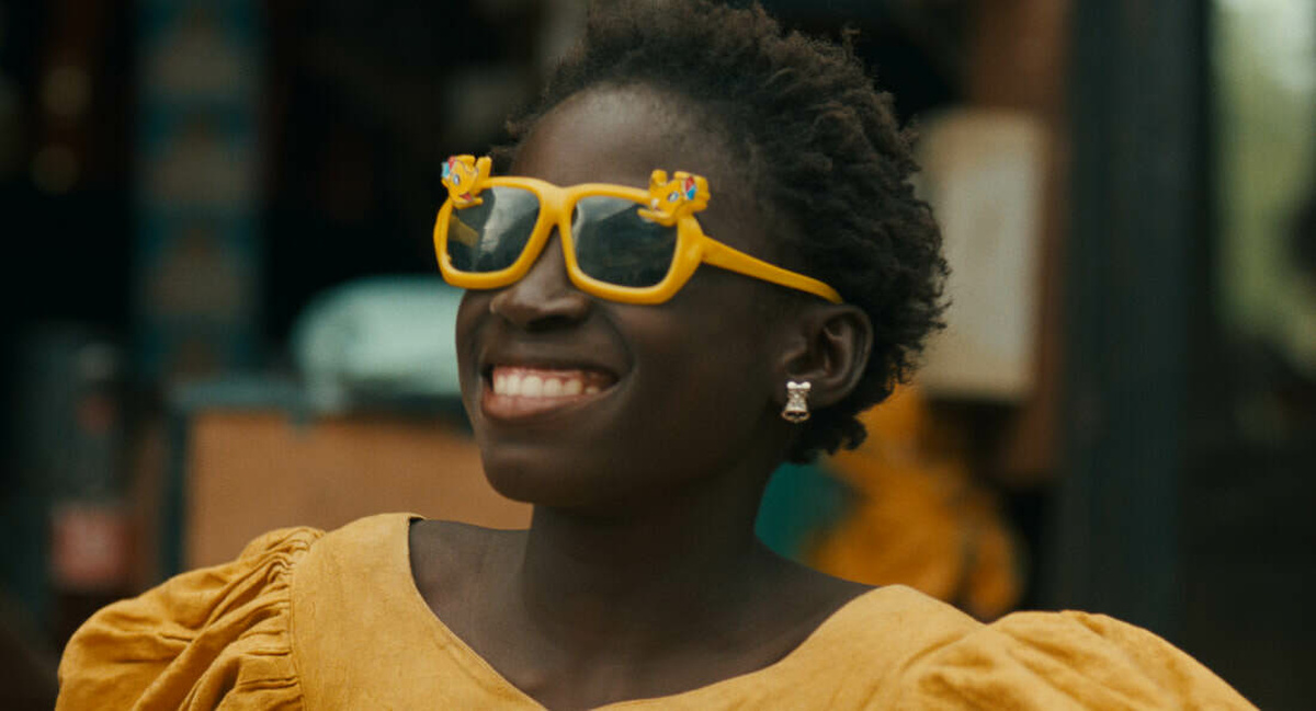 A shot of Sili smiling wearing a yellow dress and bright yellow sunglasses.