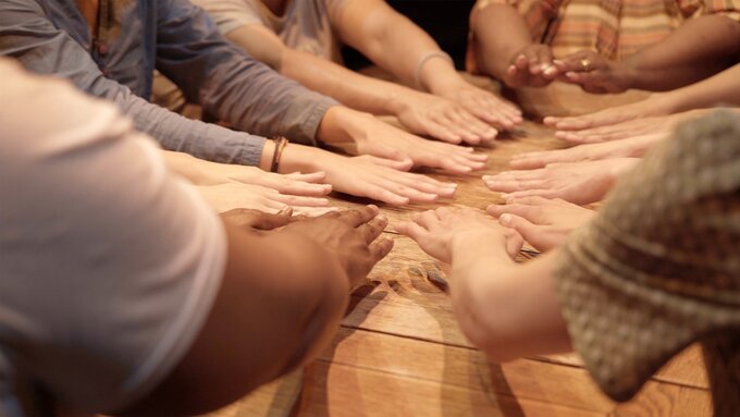Many pairs of hands hovering over a wooden table, palms facing down. In some form of communal activity.