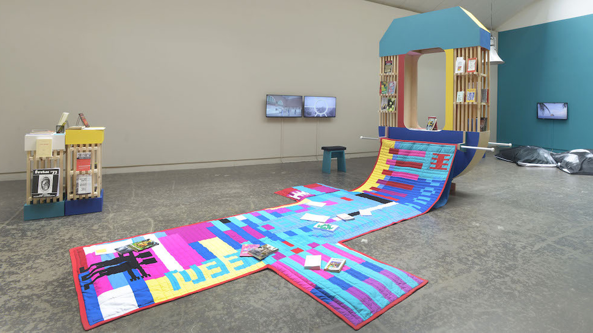 A photo of the gallery space. A large colourful geometrically patterned quilt hangs from a small mobile library.
