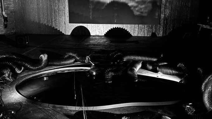 A black and white image of Umweltraum((a)). Black tubes surround a pond on the floor.