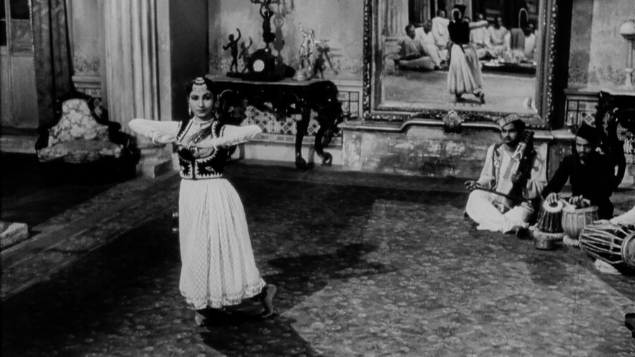 A Bengali dancer performs in an opulent room, surrounded by musicians. We can see her audience reflected in a mirror.