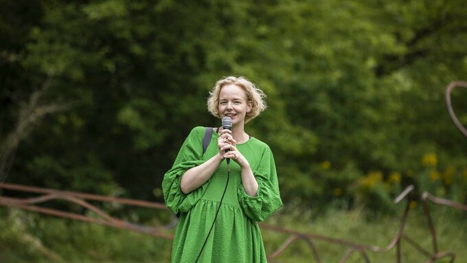 A white blonde haired woman stood holding a microphone in a field, surrounded by greenery.