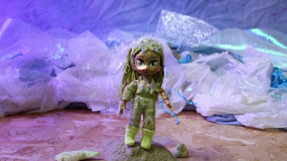 A Bratz-like doll with green hair stands on a space-like rock. Behind a sci-fi vista is made of bunched up materials.