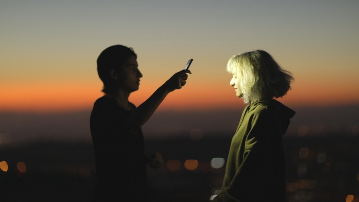 Two people stand against a sunset. One holds their phone above the other, lighting them up with the phone torchlight.