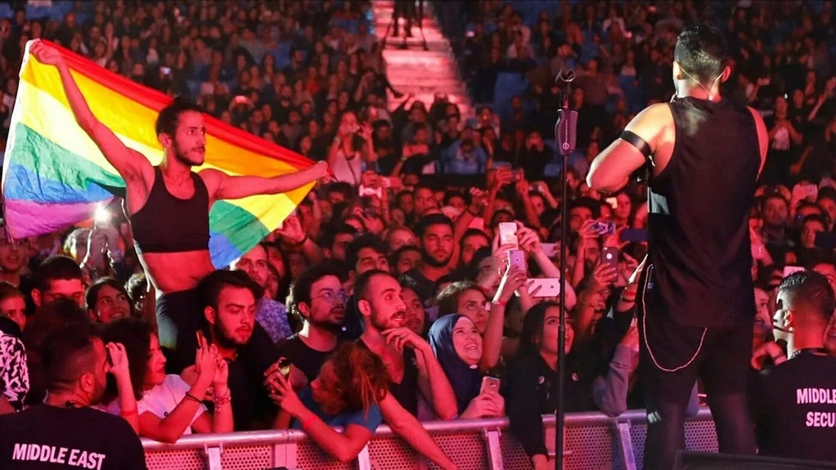 A performer stood on stage facing a concert crowd. In the crowd is a person on someones shoulders with a pride flag.