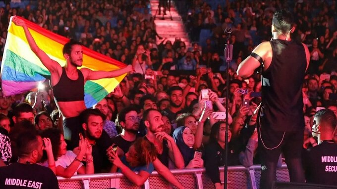 A performer stood on stage facing a concert crowd. In the crowd is a person on someones shoulders with a pride flag.