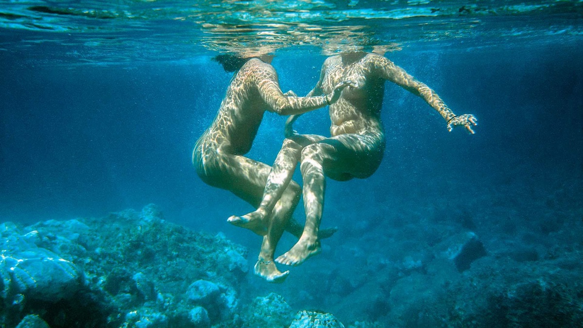 Two naked bodies float underwater. Amongst their feet is a rocky seabed.