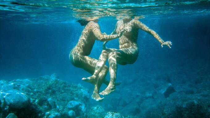 Two naked bodies float underwater. Amongst their feet is a rocky seabed.