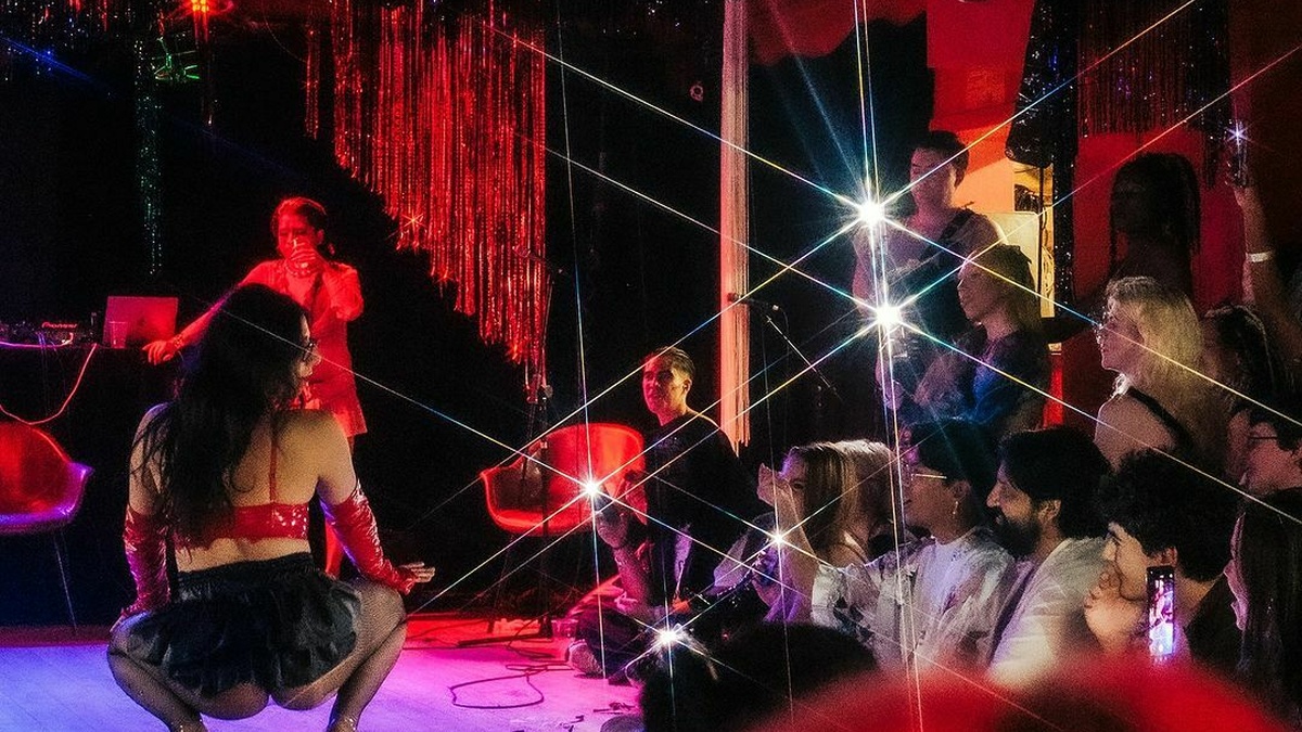 A performer crouches on stage in a performance to a live crowd. Lit with red lighting and disco ball reflections.