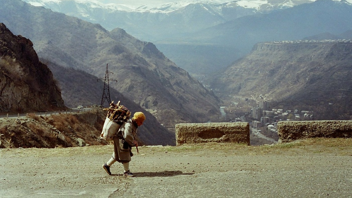 A hunched man carrying firewood along a road. Behind him is a vast mountainous landscape.