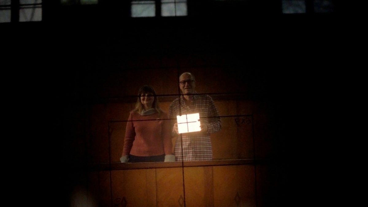 A man and a woman stood behind a frame of some kind. The scene is dark, illuminated by a light box the man holds.