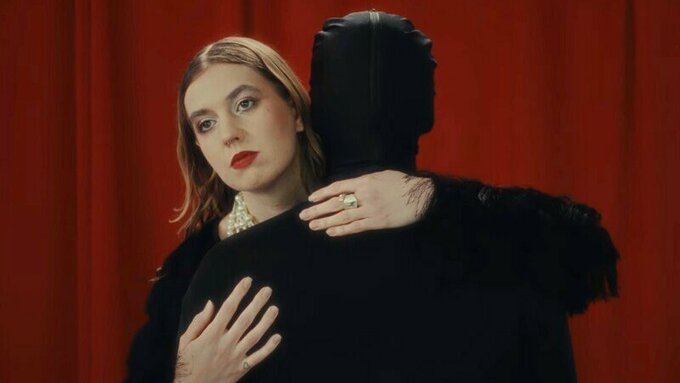 A woman wearing red lipstick hugs a black clothed figure.