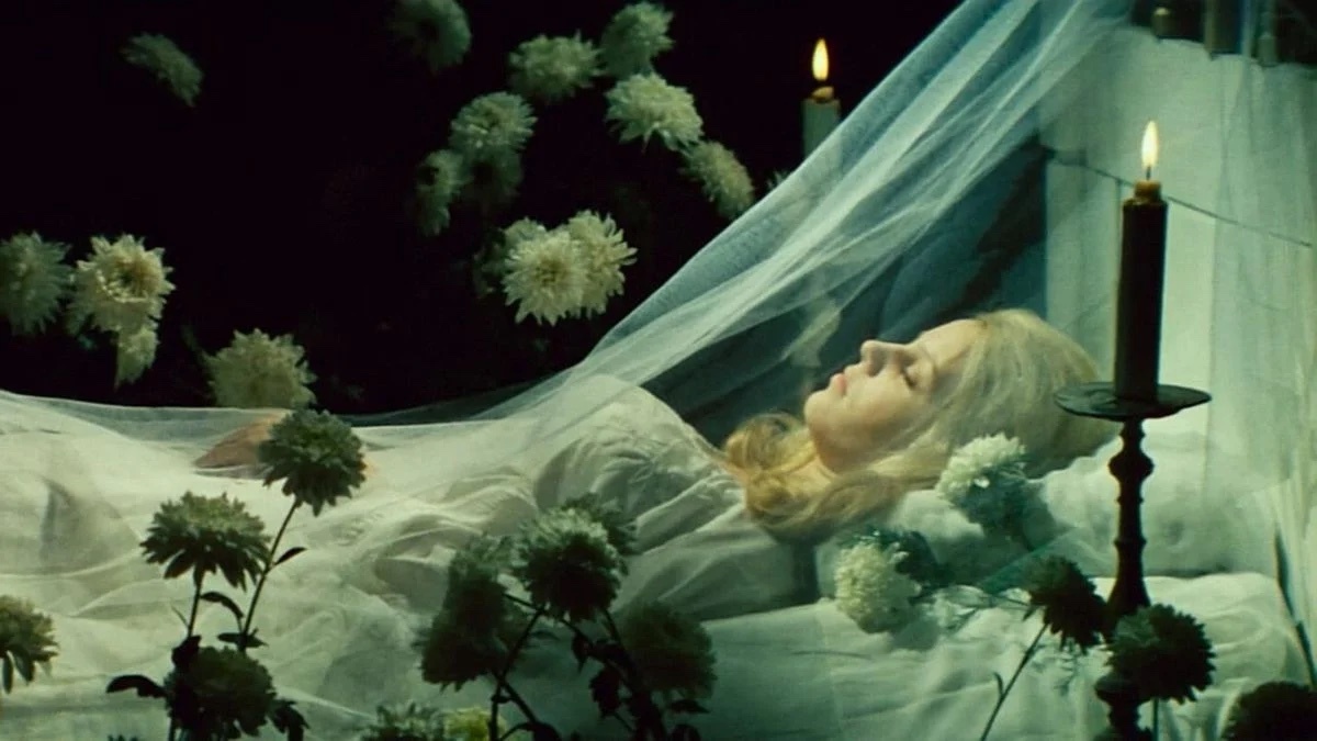 A woman with bleach blonde hair laying in a bed asleep. She is shrouded in a white veil and surrounded by white flowers.