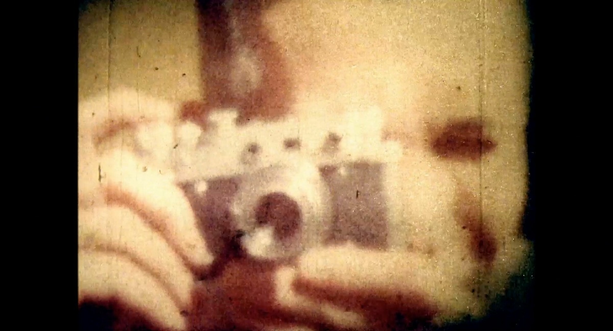 A grainy still of a person pointing a film camera at the viewer.