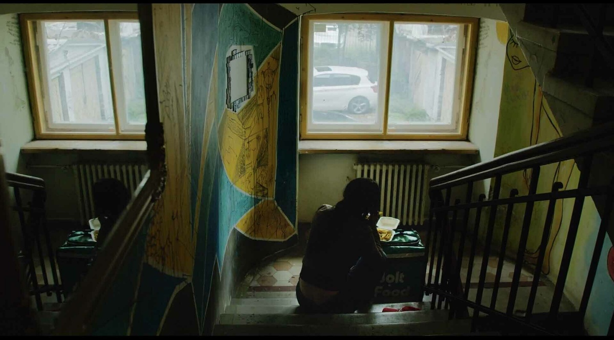 A still of a woman crouched in a stairwell beside an opened food delivery backpack.