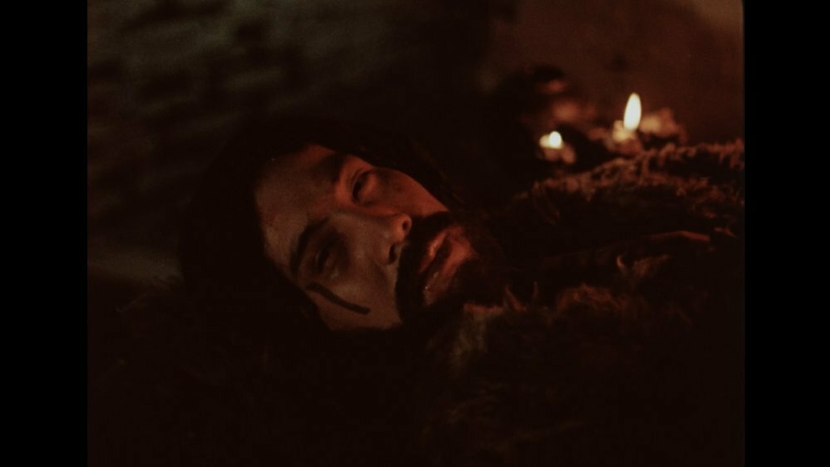 A man with a thick black moustache laying in a dark place only lit by a fire.