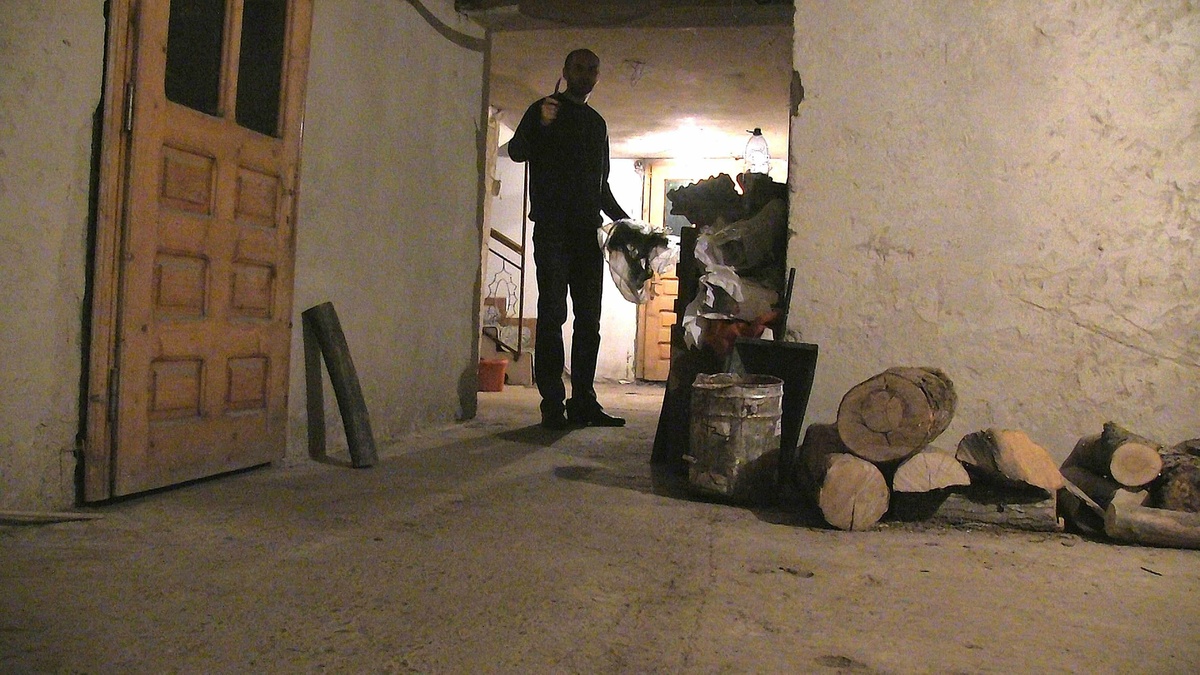 A scene in a cellar: a threatening man stood at the bottom of the stairs points a knife towards the camera.