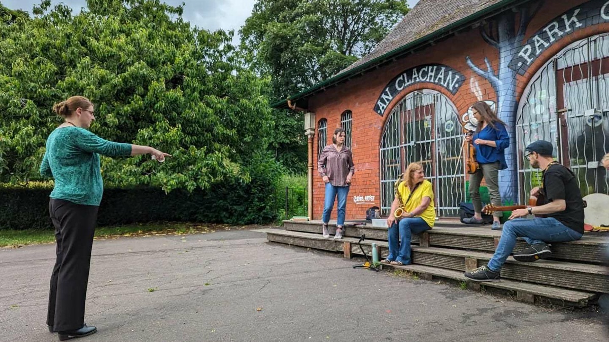 In a park in front of the 'An Clachan' building a person points commandingly at 4 figures, who hold musical instruments.