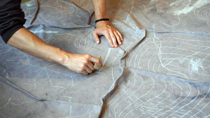 A person’s arms  reaching across a grey piece of fabric and using a tool to create marks and drawings on the fabric.