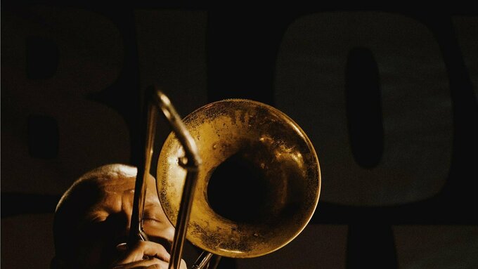 A photo portrait of a man playing the trombone in a dark space.